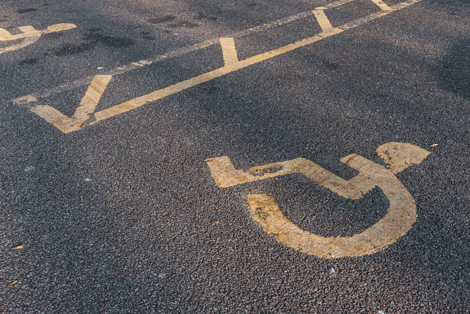 Close-up view of a disabled parking space marking on an asphalt driveway, featuring a yellow wheelchair symbol and accompanying directional lines. The painted lines are slightly worn and faded, indicating regular use. The textured asphalt surface shows small stones and roughness typical of parking areas. In the background, a segment of another lane marking is visible. This scene is outdoors, with natural lighting highlighting the surface details. The image relates to house removals and moving services offered by Man With a Van Cranford, emphasizing logistics and parking considerations during home relocation or furniture transport, especially near residential flats on Staines Road in Cranford.