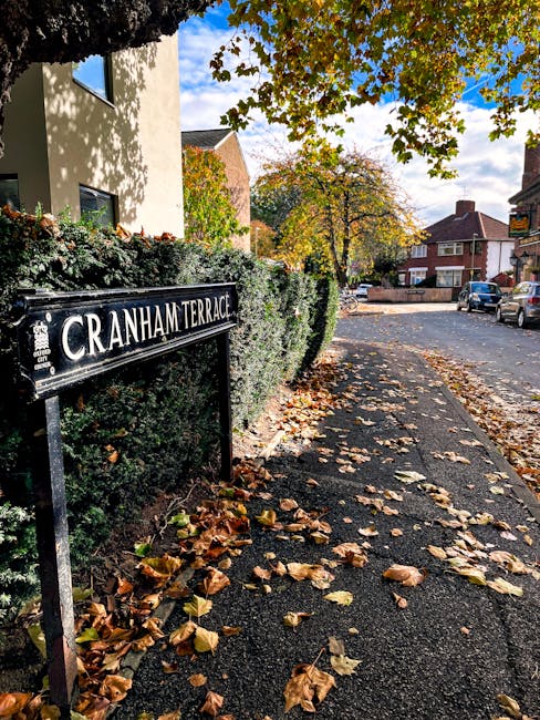 A photograph of a residential street scene showing a black and white sign reading 'CRANHAM TERRACE' attached to a metal post in front of a bushy hedge, with fallen autumn leaves scattered on the pavement. The pavement runs alongside a cream-colored building with windows, partially shaded by overhanging trees with green and yellow leaves, indicating the autumn season. In the background, there are additional residential buildings with brick facades and pitched roofs, parked cars on the street, and a blue sky with some clouds. This outdoor scene is indicative of a typical area where house removals and furniture transport services, such as those offered by Man With a Van Cranford, may be involved in home relocation and packing processes.