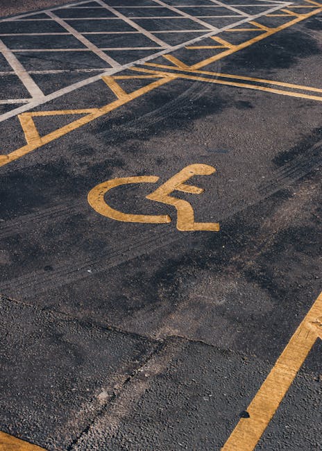 Close-up view of a disabled parking space marking on an asphalt driveway, featuring a yellow wheelchair symbol and accompanying directional lines. The painted lines are slightly worn and faded, indicating regular use. The textured asphalt surface shows small stones and roughness typical of parking areas. In the background, a segment of another lane marking is visible. This scene is outdoors, with natural lighting highlighting the surface details. The image relates to house removals and moving services offered by Man With a Van Cranford, emphasizing logistics and parking considerations during home relocation or furniture transport, especially near residential flats on Staines Road in Cranford.