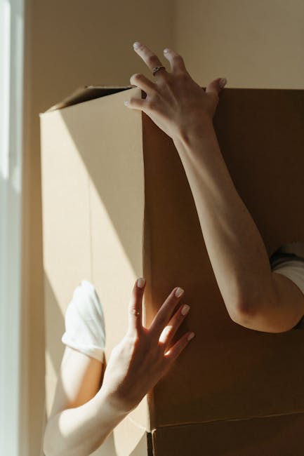 Close-up of a person's arms and hands as they lift and carry a large cardboard moving box during a home relocation. The individual's right hand grips the top edge of the box, while the left hand supports the side, with the person's torso and part of their white t-shirt visible beneath the box. The scene is set indoors near a doorway, with natural light illuminating the scene and casting shadows on the beige walls and door frame. The box appears sturdy, made of brown corrugated cardboard, suitable for packing household items during furniture transport. Inside the property, the environment suggests an organized packing process typical of professional removals, such as those offered by Man With a Van Cranford, who specialise in removals and moving logistics. Visible elements emphasize the manual handling involved in loading and packing, essential phases in a house removal or home relocation service.