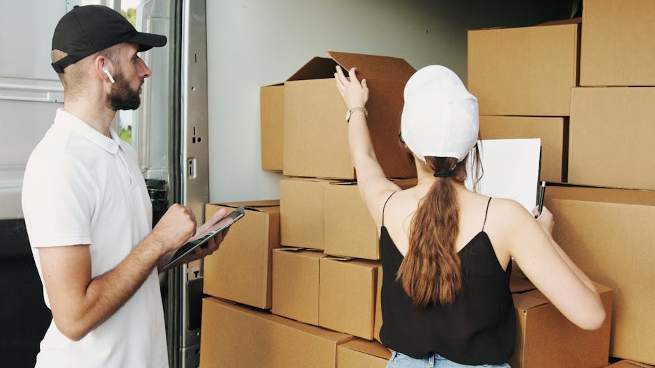 A man and a woman are inside a property near an open doorway, where multiple stacked cardboard boxes are visible. The woman, wearing a white cap and a black sleeveless top, is reaching up to adjust or inspect a box on the top of the stack while holding a clipboard in her other hand. The man, dressed in a white t-shirt and a black cap, stands nearby holding a tablet, observing the woman or possibly taking notes. The scene takes place during a home relocation process, with the boxes wrapped in cardboard and positioned on the floor close to the doorway, suggesting they are preparing for loading or unloading. Nearby, a vehicle, likely a van used for furniture transport and packing and moving, is partially visible with its door open, indicating ongoing packing or unpacking activities. The environment is well-lit with natural light, and the focus is on the careful handling of belongings as part of a professional removals service, exemplified by Man With a Van Cranford.