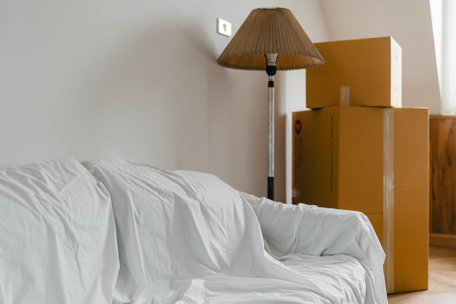 Close-up of a person's arms and hands as they lift and carry a large cardboard moving box during a home relocation. The individual's right hand grips the top edge of the box, while the left hand supports the side, with the person's torso and part of their white t-shirt visible beneath the box. The scene is set indoors near a doorway, with natural light illuminating the scene and casting shadows on the beige walls and door frame. The box appears sturdy, made of brown corrugated cardboard, suitable for packing household items during furniture transport. Inside the property, the environment suggests an organized packing process typical of professional removals, such as those offered by Man With a Van Cranford, who specialise in removals and moving logistics. Visible elements emphasize the manual handling involved in loading and packing, essential phases in a house removal or home relocation service.