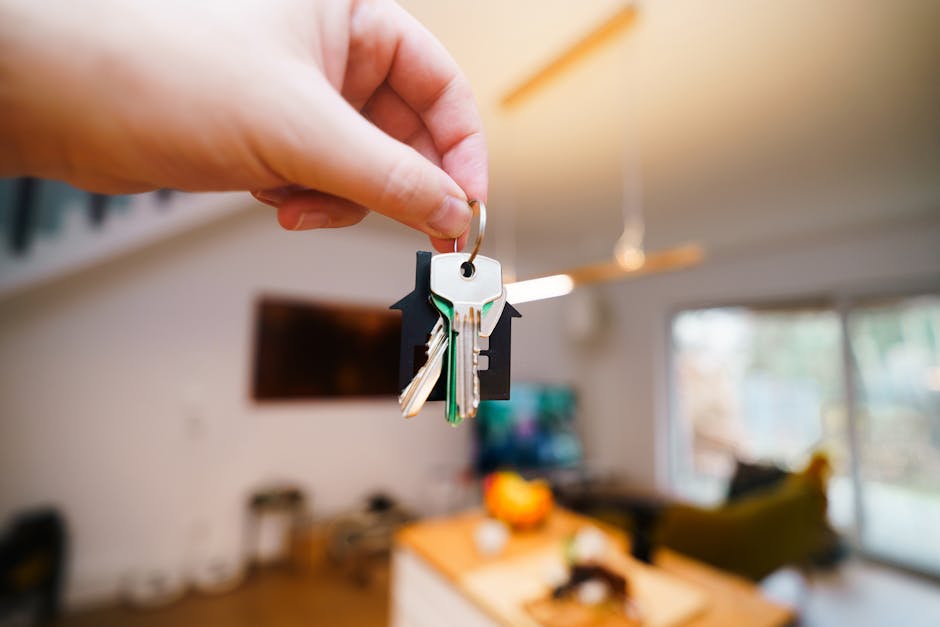 A close-up of a person's hand holding a keyring with three keys, including a silver key and a smaller green key, over a living room with modern furnishings. The background features a television mounted on the wall, a sofa with cushions, a coffee table with decorative items, and large windows allowing natural light to illuminate the space. The keys are likely used for house entry during home relocation or packing and moving activities. This scene correlates with furniture transport and moving logistics typical of a residential property, as part of professional removals services provided by Man With a Van Cranford, which assists in avoiding common house removals mistakes for Cranford landlords.