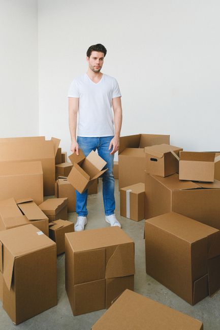 A young man wearing a white t-shirt and blue jeans stands amidst an array of brown cardboard moving boxes of various sizes, some open and partially filled, inside a plain white room with a concrete floor. He holds an open box in his right hand while looking slightly to the left. The scene depicts the packing process during a home relocation, with boxes prepared for transport. Behind him are more stacked boxes on the floor, with some positioned near the doorway, indicating an active packing or unpacking phase. The lighting in the room is bright and natural, highlighting the moving materials and the workspace for furniture transport and packing. This image illustrates the process of organizing and preparing belongings for a house move with professional removals services, exemplifying careful preparation and logistics involved in house removals or moving projects. Man With a Van Cranford offers assistance with such moving procedures, focusing on efficient packing and loading during furniture transport.