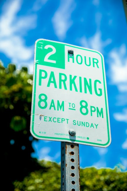 A close-up image of a green and white parking restriction sign mounted on a metal pole outdoors, with a clear blue sky and green foliage in the background. The sign indicates a two-hour parking limit from 8 am to 8 pm, Monday to Saturday, with exemption for Sundays. The sign is attached to the pole with two bolts and is situated near a residential or urban area, relevant to house removals and moving services provided by Man With a Van Cranford. The environment suggests an accessible, well-maintained parking area suitable for loading and unloading furniture during home relocation or removal services, supporting transport logistics for local house moves in Cranford.