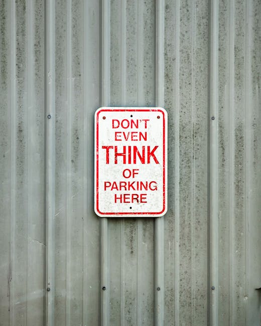 A rectangular metal sign with a white background and red border is mounted on a vertically corrugated metal wall. The sign displays the message 'DON'T EVEN THINK OF PARKING HERE' in bold red capital letters, with the words 'THINK' and 'HERE' emphasized by larger font size. The wall has a weathered appearance with visible stains and slight discoloration. The sign's positioning suggests it is situated outdoors, possibly near an entrance or loading area, which is relevant for house removals and furniture transport in Cranford. This image relates to parking regulations for removal vehicles during home relocation processes, as managed by Man With a Van Cranford, to ensure compliance with local parking rules during the loading and unloading phases of a move.
