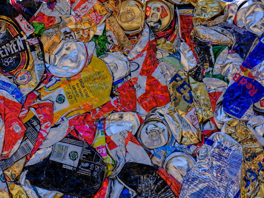 A close-up view of crumpled and flattened aluminum beverage cans, including brands such as Pepsi and other soft drinks, scattered and overlapped on a surface. The cans display various colors like silver, blue, red, yellow, and black, with some labels partially visible. The metallic surfaces show dents, scratches, and creases from prior use. This image depicts materials commonly involved in recycling and waste sorting, relevant to house removals and packing processes handled by Man With a Van Cranford. The cans are likely prepared for recycling or disposal during a home relocation or waste management task, with no other furniture or packaging materials present in the image, emphasizing the need for proper waste separation during moving services.