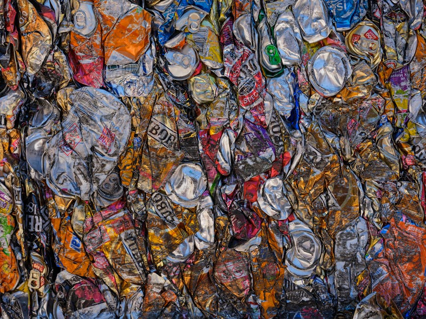 A large pile of crumpled aluminium cans, mostly silver and metallic, with some orange, red, blue, green, and purple labels visible, gathered together on a surface. The cans are tightly packed, with many overlapping each other, creating a dense, textured appearance. The image appears to be taken from above, showcasing the volume of recyclable waste, possibly as part of a packing or disposal process during a house removal or relocation. This visual relates to waste sorting and recycling procedures, which are relevant to moving and home relocation services offered by Man With a Van Cranford, especially in contexts such as house clearance or reusable material handling. The environment suggests an indoor setting, with the cans possibly stored temporarily before collection or disposal, highlighting the importance of planning for waste management during a home move.
