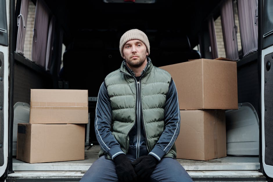 A man with a beard and serious expression sitting on the edge of an open van trunk during a house removal, surrounded by cardboard boxes of various sizes, some wrapped in plastic or packing paper, ins