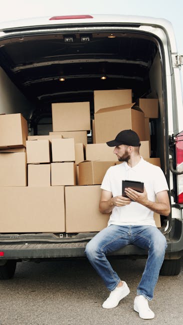A man with a beard and serious expression sitting on the edge of an open van trunk during a house removal, surrounded by cardboard boxes of various sizes, some wrapped in plastic or packing paper, ins