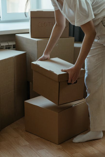 A person wearing a white shirt, beige trousers, and white socks is handling a cardboard box during a home relocation process, with several other packed boxes stacked nearby. The individual's hands are gripping the sides of the box as they prepare to lift or move it. The boxes are uniformly sealed with packing tape, some labeled with handwritten text, such as 'clothes,' indicating contents. They are positioned on a light wooden floor inside a room with natural light coming through large windows. Behind the person, there is a stack of additional boxes and packing materials, suggesting an active packing and moving operation. In the background, part of a sofa and curtains are visible, implying a residential setting. This scene exemplifies the packing and furniture transport actions involved in a professional removals service, such as those offered by Man With a Van Cranford, during house removals or relocations in Cranford.