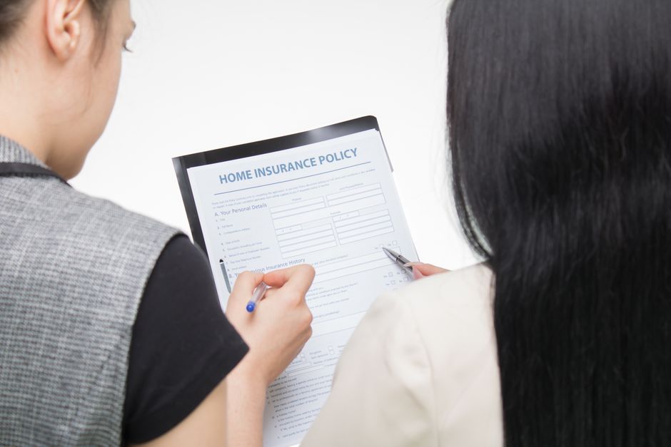 Two women are seated indoors during a home relocation process, reviewing a document titled 'Home Insurance Policy' that is held on a clipboard by one woman while the other points at it with a pen. The woman on the left has a gray vest with black sleeves, and the woman on the right has long dark hair and is wearing a light-colored blazer. The environment is well-lit, and the focus is on the paperwork related to moving and insurance coverage. This scene illustrates aspects of packing, moving logistics, or insurance preparations associated with house removals, as seen with the focus on documentation and partnership. Man With a Van Cranford provides professional removals services, including packing and moving coordination, supporting home relocations efficiently, as reflected by the focus on essential paperwork in this image.