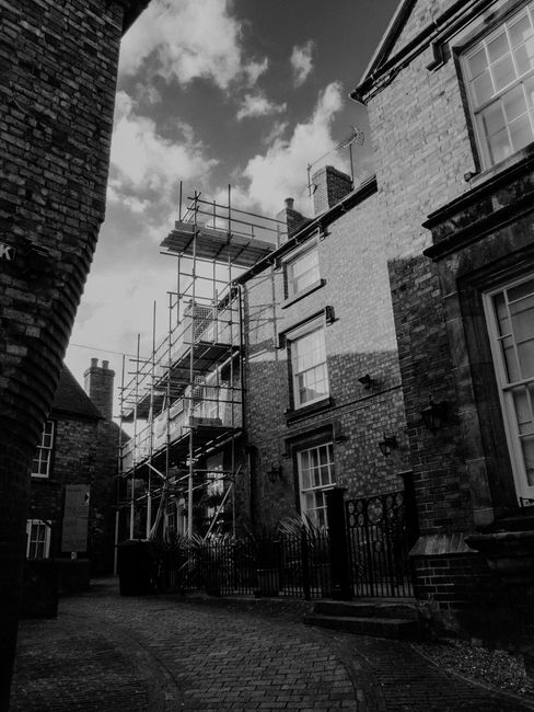 Black and white photograph showing the exterior of a residential property during a home relocation process, with scaffolding erected along the side of the building. The scaffold comprises metal tubes