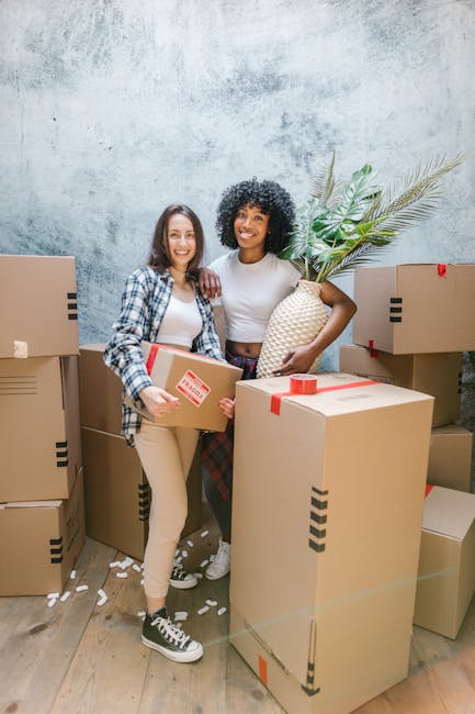 Two women involved in a home relocation arranged by Man With a Van Cranford are standing among stacked moving boxes of various sizes, all sealed with red packing tape. The woman on the left is wearing a checkered shirt over a white top, beige pants, and black and white sneakers, holding a medium-sized box. The woman on the right, dressed in a white top and patterned trousers, is smiling while holding a large, woven vase with tall, leafy green plants. The scene takes place indoors, with a textured grey wall in the background and a wooden floor beneath their feet. Several packing materials, such as crumpled paper, are scattered on the floor around them, indicating an active packing and moving process. This image illustrates the packing stage of a furniture transport or house removals service, highlighting the careful handling of furniture and boxes by professionals like Man With a Van Cranford during home relocation preparations.