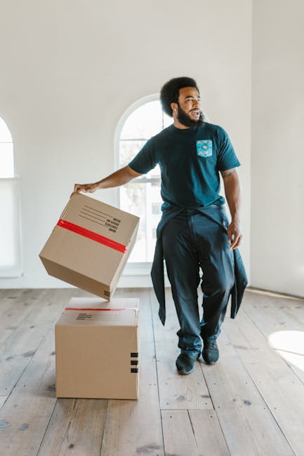 A man with dark curly hair and a beard, wearing a navy blue t-shirt with a light blue logo and dark trousers, is inside a bright, empty room with white walls and a large arched window that allows natural light to illuminate the space. He is in the process of packing or unpacking, holding a medium-sized cardboard box reinforced with red tape in his right hand, while another closed box rests on the wooden floor nearby. The room appears to be in a house prepared for moving, with the man possibly part of a professional removal service such as Man With a Van Cranford, who are involved in home relocation or furniture transport. The scene captures aspects of packing and moving logistics, highlighting materials like cardboard boxes and the natural daylight that emphasizes a calm, organized environment for house removals.
