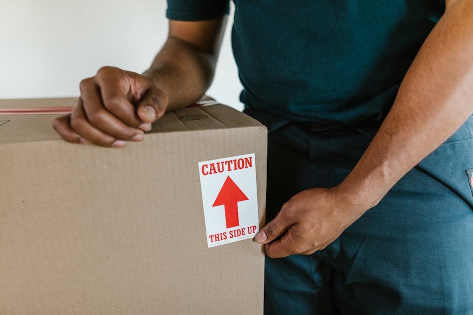 A person wearing a dark green shirt and blue trousers is holding a large cardboard moving box with both hands, ensuring proper grip during a home relocation process. The box is sealed with packing tape, and a white label with red text and an upward arrow reads 'Caution This Side Up,' indicating careful handling and proper orientation. The setting appears to be indoors, near a doorway or transition area, with natural lighting illuminating the scene. This image reflects the careful packing, handling, and loading process involved in furniture transport and home removals, which are services provided by Man With a Van Cranford, supporting clients during their house moves along Bath Road in Cranford.