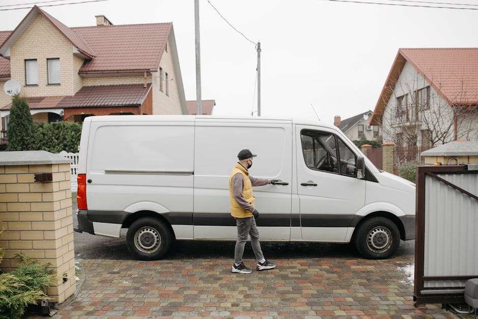 A man wearing a black cap, glasses, a yellow and grey vest, grey pants, and sneakers is standing outside a residential property, opening the sliding door of a white delivery van parked on a paved driveway. The van is positioned parallel to the house, with the driver's side facing the camera. The scene is set in a suburban neighbourhood with detached houses that have pitched, red-tiled roofs and light-colored walls, some with dormer windows and chimneys. A brick wall and a garden fence surround the property, and nearby there is a small, leafy green shrub and a garbage bin. The overcast sky indicates cloudy weather, and the lighting is diffuse, typical of an overcast day. This image depicts a home relocation or furniture transport process, with [COMPANY_NAME], Man With a Van Cranford, providing removals and moving services, involving loading or unloading items from the van in a typical moving logistics setting.