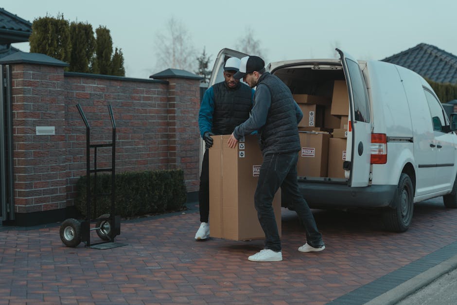 A smiling man wearing a white polo shirt and black cap is standing outdoors beside a van during a home relocation process. He is holding a medium-sized cardboard box, with several other packed boxes stacked inside the open rear door of the van. The boxes are made of cardboard and are sealed with packing tape. The van is positioned on a paved surface near a grassy area with trees and water visible in the background. Inside the van, additional boxes and packing materials, such as plastic wraps or protective padding, may be present, indicating an ongoing packing and loading process. The scene captures the logistics of furniture transport and packing during a professional move, with a focus on the careful handling of household items. [COMPANY_NAME], an experienced removals service, is involved in this home relocation effort, demonstrating the typical steps of loading boxes into a van for efficient transportation to a new property.
