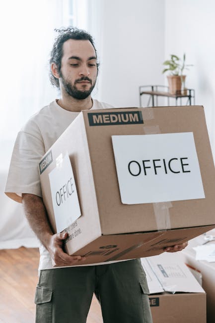 A man with dark, curly hair and beard, wearing a light-colored T-shirt and khaki cargo pants, is indoors carrying a large cardboard box labeled 'OFFICE' in black text along with a smaller label indicating 'MEDIUM' size. The background shows a bright room with white walls, wooden flooring, and minimal furnishings, including a window with natural light, a small table with a potted plant, and some packing materials scattered on the floor. The scene depicts the process of packing or unpacking during a house or office relocation, which is part of the services provided by Man With a Van Cranford for office-to-office moves and home removals.