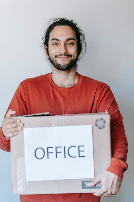 A man with dark, curly hair and a beard is standing indoors against a plain light grey wall, smiling softly while holding a cardboard box labeled 'OFFICE' with both hands. The box is sealed with clear packing tape and features a recycling symbol in the upper right corner. The man is dressed in a red long-sleeve shirt, and his relaxed posture suggests he is preparing for or engaged in office relocation or packing activities. The image emphasizes professional packing and moving processes, fitting with house and office removals services offered by Man With a Van Cranford, focusing on furniture transport and logistics during a home or business relocation.