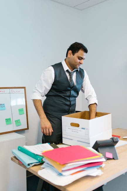 A man with dark, curly hair and beard, wearing a light-colored T-shirt and khaki cargo pants, is indoors carrying a large cardboard box labeled 'OFFICE' in black text along with a smaller label indicating 'MEDIUM' size. The background shows a bright room with white walls, wooden flooring, and minimal furnishings, including a window with natural light, a small table with a potted plant, and some packing materials scattered on the floor. The scene depicts the process of packing or unpacking during a house or office relocation, which is part of the services provided by Man With a Van Cranford for office-to-office moves and home removals.